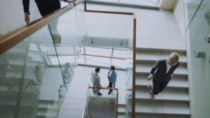 Three individuals interacting in a modern office staircase setting with glass railings and tiled steps.